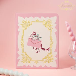 May include: Pink notebook featuring a whimsical illustration of a chipmunk holding a pink envelope with a blue bow. The design is framed by a gold floral border on a cream background, with a pink border. The Suchakk logo is in the upper right corner.