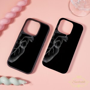May include: Two black phone cases featuring a white anatomical heart design. The cases are displayed on a pink surface, with a pink candle and a glass in the background. The cases have a Suchakk logo.