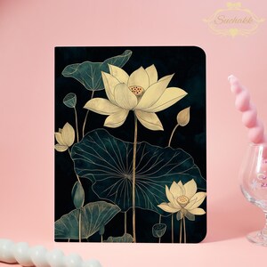 May include: A notebook with a dark background featuring a golden lotus flower design. The lotus flowers and leaves are detailed with gold outlines, creating a striking contrast against the dark backdrop. The notebook has a Suchakk logo.