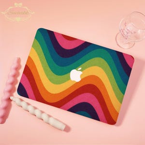 May include: A laptop case featuring a colourful rainbow wave design and a white Apple logo. The case is on a pink surface, with a glass and two decorative candles. The Suchakk logo is in the top left corner.