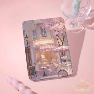 Op de afbeelding: Een tablethoes met een pastelkleurige illustratie van een Parijse bakkerij. De afbeelding toont een roze en witte gestreepte luifel, een bloemenkar en een kersenboom. Het bakkerijbord luidt "Cindae & Sainage". Een glas met tekst staat op de achtergrond.