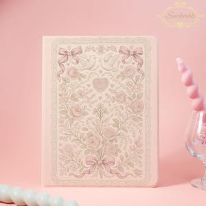 May include: A rectangular notebook with a light pink cover featuring a detailed illustration of two white doves, a central heart, roses, foliage, and two bows. The design is framed by a lace-like border. The text Suchakk appears in the top right corner.