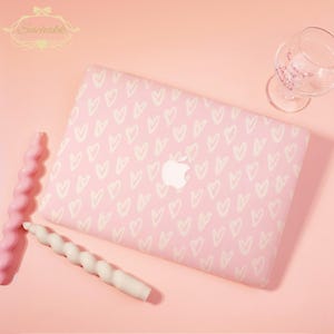 May include: Pink laptop case with a white heart pattern. The case has the Apple logo in the center. A pink and white twisted candle and a wine glass are also in the image. The background is a solid pink.