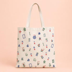May include: Off-white canvas tote bag with a repeating pattern of Mahjong tiles in red, green, and blue. The bag has a rectangular shape with a matching canvas handle. The design includes Chinese characters and various tile illustrations.