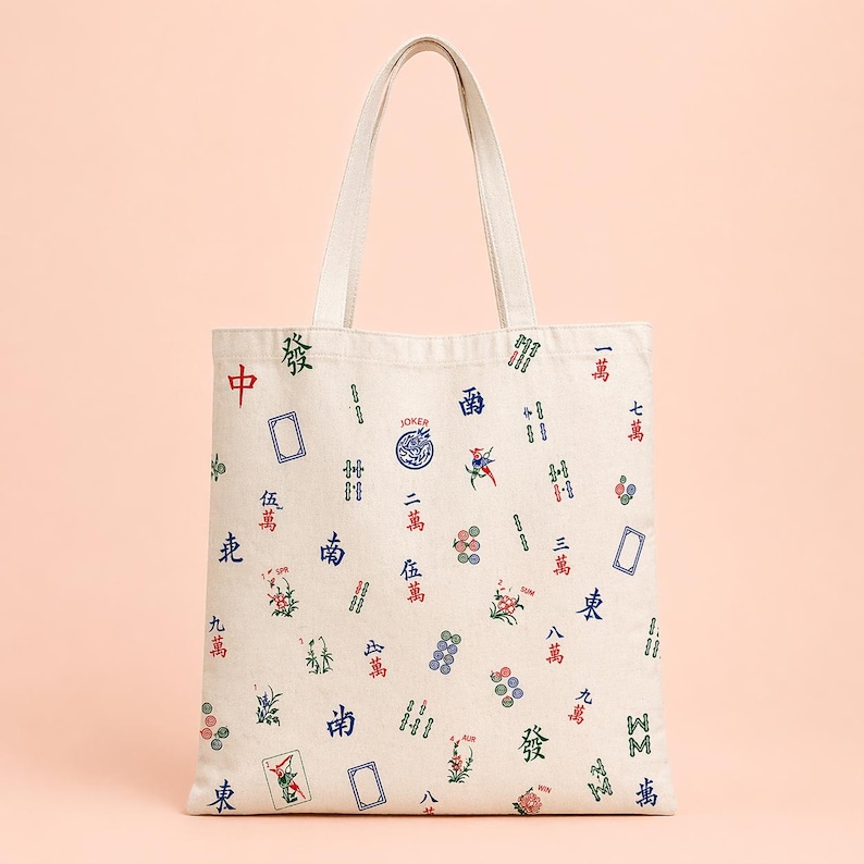 May include: Off-white canvas tote bag with a repeating pattern of Mahjong tiles in red, green, and blue. The bag has a rectangular shape with a matching canvas handle. The design includes Chinese characters and various tile illustrations.
