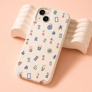 May include: A white iPhone case featuring a colorful Mahjong tile design. The case has a cream-colored background with various symbols and Chinese characters in red, blue, and green. The case has a dual-camera cutout.