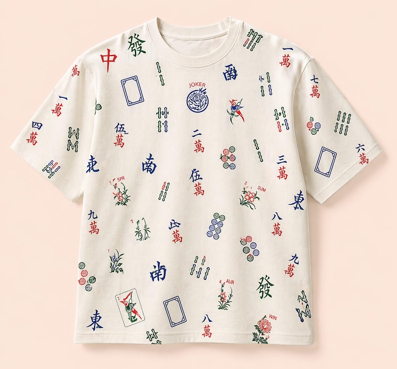 May include: Cream-colored t-shirt with a round neck, featuring a pattern of Mahjong tiles and Chinese characters in red, blue, and green. The word "JOKER" is printed on the shirt. The shirt has short sleeves.