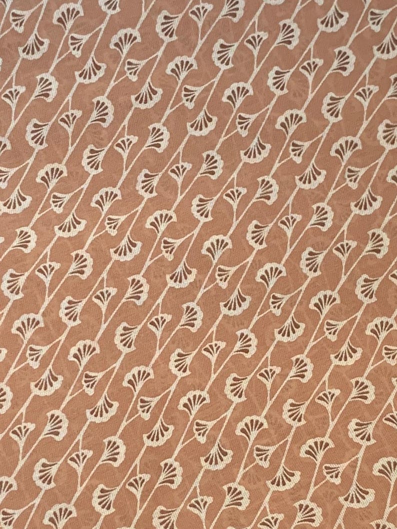 K&ouml;nnte beinhalten: Ein Stoff mit einem sich wiederholenden Muster aus wei&szlig;en, f&auml;cherf&ouml;rmigen Ginkgobl&auml;ttern, die in diagonalen Linien auf einem einfarbigen, terrakottafarbenen Hintergrund angeordnet sind. Das Design erzeugt eine konsistente, strukturierte Textur.