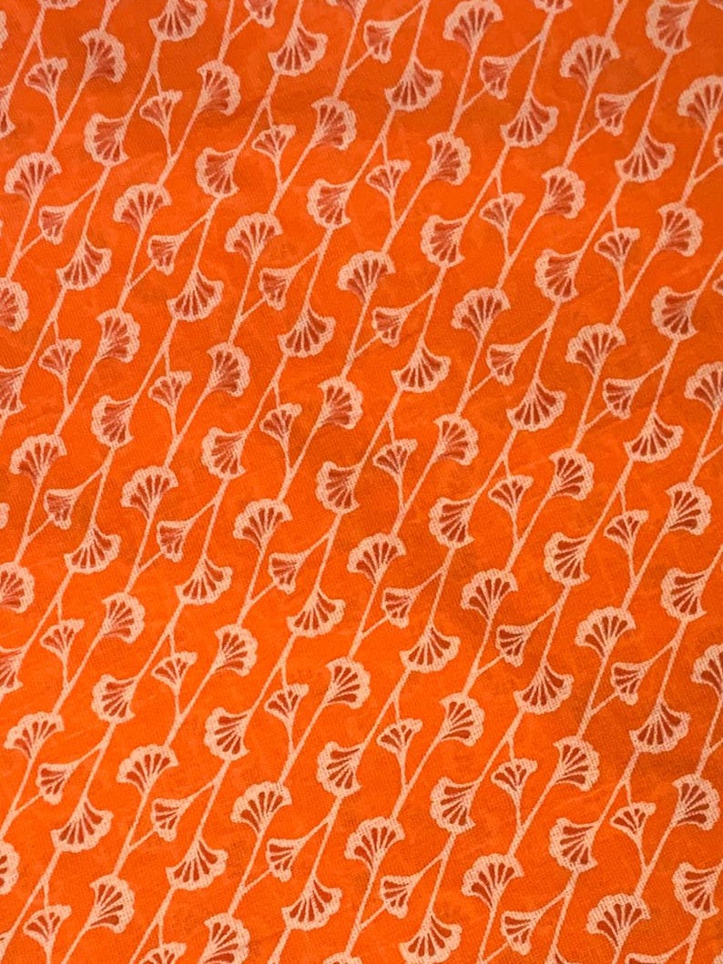 K&ouml;nnte beinhalten: Ein leuchtend orangefarbener Stoff mit einem sich wiederholenden Muster aus wei&szlig;en, diagonalen Linien. Jede Linie ist mit kleinen, f&auml;cherf&ouml;rmigen Blattmotiven verziert, die in einer konsistenten, rhythmischen Abfolge &uuml;ber die gesamte Textiloberfl&auml;che angeordnet sind.