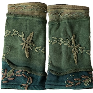 Op de afbeelding: Een paar groene armwarmers met geborduurde bloemmotieven. De armwarmers hebben een donkergroene basis met lichtgroene en bruine borduursels. De boven- en onderranden hebben een donkergroene rand.