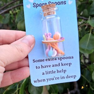 May include: A small glass bottle filled with miniature colourful spoons, secured with a rubber band, and a cork stopper. The bottle is on a blue card with the text "Spare Spoons" and "Some extra spoons to have and keep a little help when you're in deep."