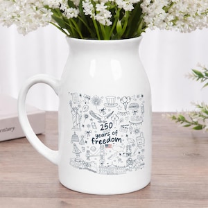May include: A white ceramic pitcher with a handle, filled with white flowers. The pitcher features a black and white illustration with the text "250 years of freedom" and various American icons.