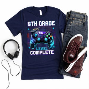 May include: Navy blue t-shirt with the text "8th Grade Level Complete" and a graphic of a video game controller with a graduation cap. The image also includes a pair of jeans, burgundy sneakers, and black headphones.