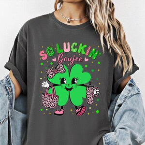 May include: Dark gray t-shirt with a St. Patrick's Day graphic. The graphic features a smiling green four-leaf clover character with a pink bow, holding a leopard print tote bag and a matching tumbler. Text reads "So Luckin' Boujee".