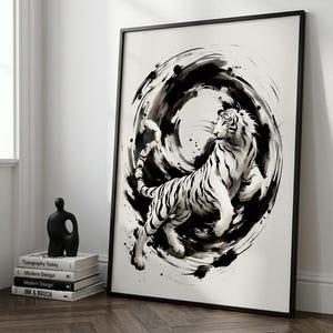 Può includere: Una stampa d'arte incorniciata che raffigura un'illustrazione a inchiostro bianco e nero di una tigre in movimento su uno sfondo circolare vorticoso. L'opera è esposta in una sottile cornice nera, appoggiata a una parete accanto a una pila di quattro libri.