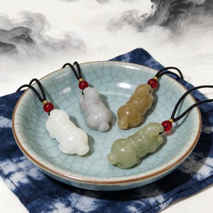 May include: Four jade pendants in varying colors: white, gray, tan, and green. Each pendant is carved in the shape of a small animal and strung on a black cord with a red bead accent. The pendants are displayed in a light blue ceramic bowl.