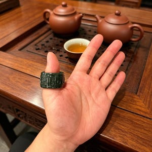 May include: A dark green jade ring with carved characters is displayed on a hand. The ring is cylindrical and has a detailed pattern. In the background, there are two brown teapots, a teacup, and a wooden table.