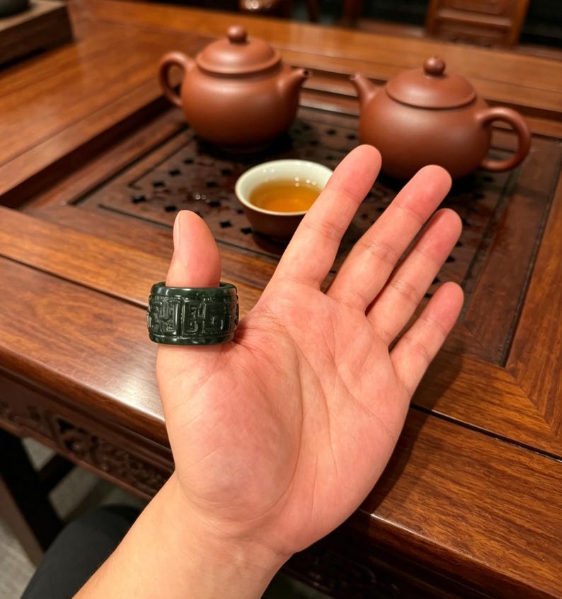May include: A dark green jade ring with carved characters is displayed on a hand. The ring is cylindrical and has a detailed pattern. In the background, there are two brown teapots, a teacup, and a wooden table.
