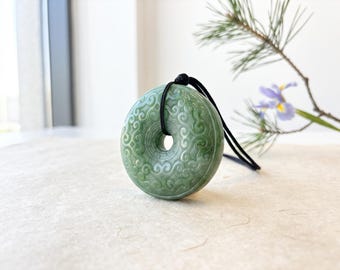 May include: A circular green jade pendant featuring an intricate carved scrollwork pattern on its surface. The pendant has a central hole and is suspended from a simple black cord, creating a balanced and detailed piece of jewellery for the neck.