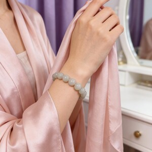 May include: A light gray beaded bracelet is worn on a person's wrist. The bracelet is made of round, smooth beads. The person is wearing a pink robe. The background includes a white vanity and a mirror.