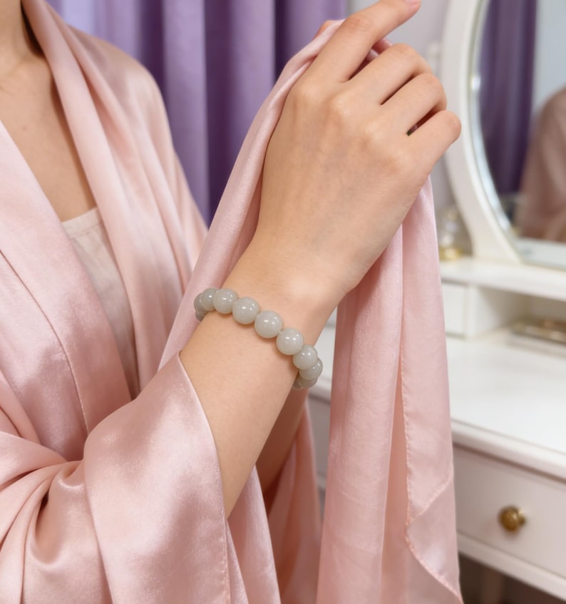 May include: A light gray beaded bracelet is worn on a person's wrist. The bracelet is made of round, smooth beads. The person is wearing a pink robe. The background includes a white vanity and a mirror.