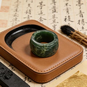 May include: A green jade ring with carved floral designs rests on a brown leather ink pad. The ink pad has a black ink well and is surrounded by calligraphy paper and a brush.