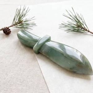 May include: A smooth, polished green stone object shaped like a double-ended blade or dagger with a central rectangular band. The stone features natural variations in color, ranging from light to dark sage green with a glossy, reflective surface finish.