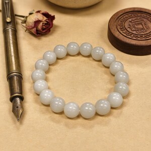 May include: A white beaded bracelet with large, round beads. The bracelet is displayed on a neutral surface, alongside a vintage brass fountain pen, a dried rose, and a wooden lid with carved details. The beads appear to be approximately 0.5 inches in diameter.