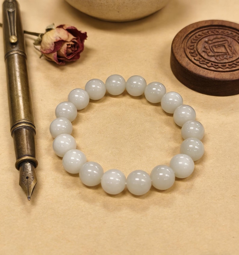 May include: A white beaded bracelet with large, round beads. The bracelet is displayed on a neutral surface, alongside a vintage brass fountain pen, a dried rose, and a wooden lid with carved details. The beads appear to be approximately 0.5 inches in diameter.