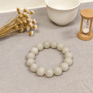 May include: A white beaded bracelet with large, round beads. The bracelet is resting on a gray surface, alongside a white teacup, dried flowers, and a small wooden hourglass.