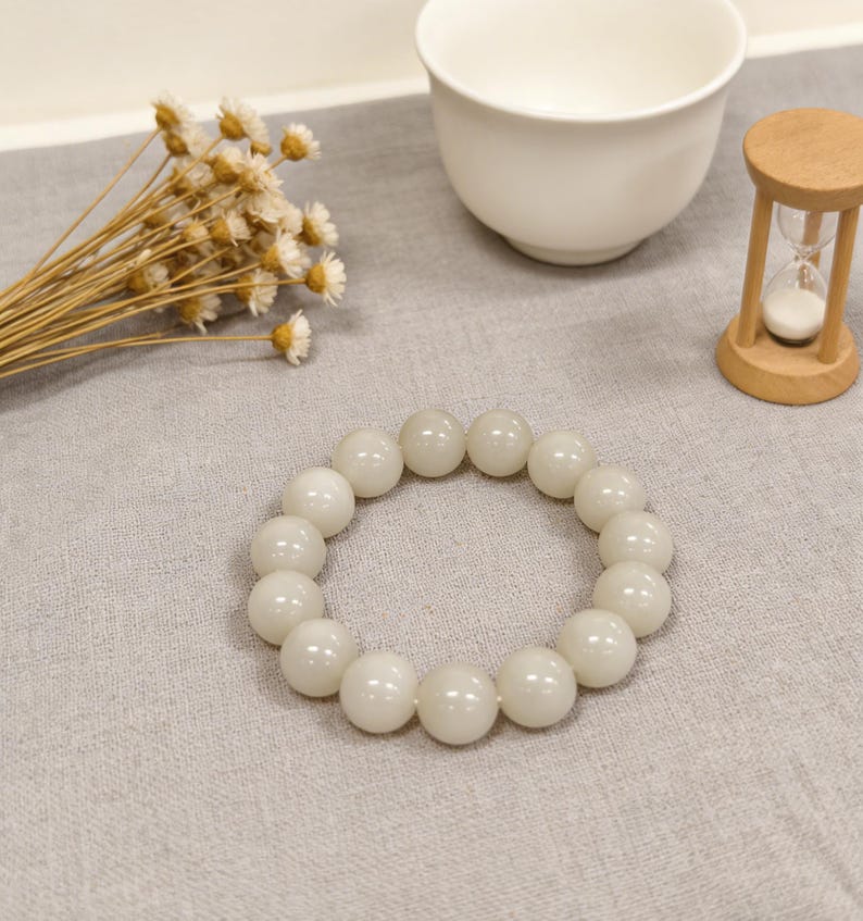 May include: A white beaded bracelet with large, round beads. The bracelet is resting on a gray surface, alongside a white teacup, dried flowers, and a small wooden hourglass.