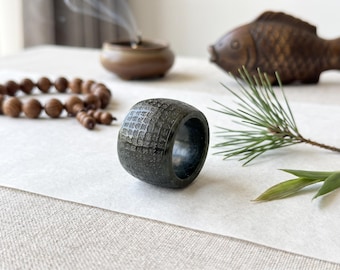 May include: A dark, cylindrical stone ring featuring a textured, grid-like pattern carved into its outer surface. The ring has a smooth, polished interior and a thick, substantial band design, showcasing a deep charcoal colour with subtle variations.
