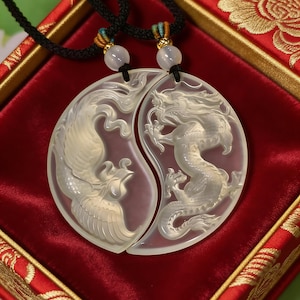 May include: Two matching pendants, each carved with a mythical creature, a phoenix and a dragon, in a yin-yang design. The pendants are light-colored and hang on black cords with decorative beads. The background is a red velvet surface.