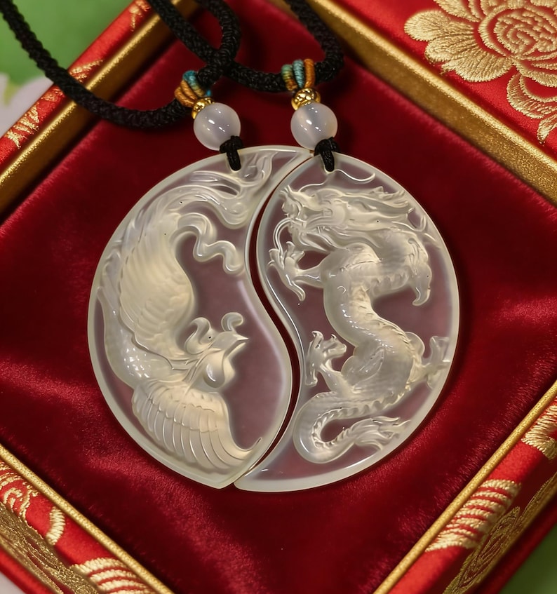 May include: Two matching pendants, each carved with a mythical creature, a phoenix and a dragon, in a yin-yang design. The pendants are light-colored and hang on black cords with decorative beads. The background is a red velvet surface.