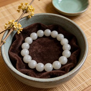 May include: A white beaded bracelet rests in a ceramic bowl lined with brown fabric. The bracelet is composed of round, opaque beads. A sprig of yellow flowers and a small green dish are in the background.