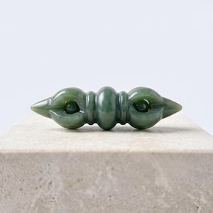 May include: A smooth, polished green jade object shaped like a double-ended vajra. The piece features a central rounded section flanked by two symmetrical, pronged ends that curve inward to meet at a pointed tip, showcasing a translucent finish.