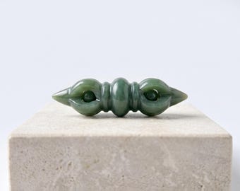 May include: A smooth, polished green jade object shaped like a double-ended vajra. The piece features a central rounded section flanked by two symmetrical, pronged ends that curve inward to meet at a pointed tip, showcasing a translucent finish.