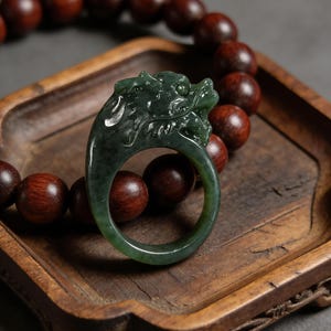 Könnte beinhalten: Dunkelgrüner Jade-Ring in Form eines Drachen. Der Ring ist vor dem Hintergrund von Holzperlen und einem Holztablett platziert. Der Drachenkopf ist detailliert mit Schuppen und Merkmalen.