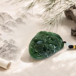 May include: A dark green jade pendant featuring an intricate relief carving of a landscape with trees and mountains. The pendant is attached to a black braided cord with a small light-colored bead and yellow thread accents near the attachment point.