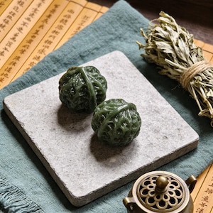 May include: Two dark green, carved stone sculptures resembling cabbages rest on a gray, square stone. The stone sits on a teal cloth, with dried herbs and a small, ornate bronze incense burner nearby. The background includes a bamboo mat.