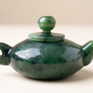 May include: A small, polished dark green stone teapot with a smooth, rounded body, a short spout, and a circular handle with a center hole. The teapot features a matching removable lid topped with a small, spherical knob, showcasing natural mottled color variations throughout the stone surface.