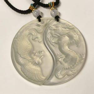 May include: A pair of light-colored pendants, each in a half-circle shape, forming a Yin and Yang symbol. One pendant features a carved dragon, the other a phoenix. The pendants are connected to black cords with decorative beads.