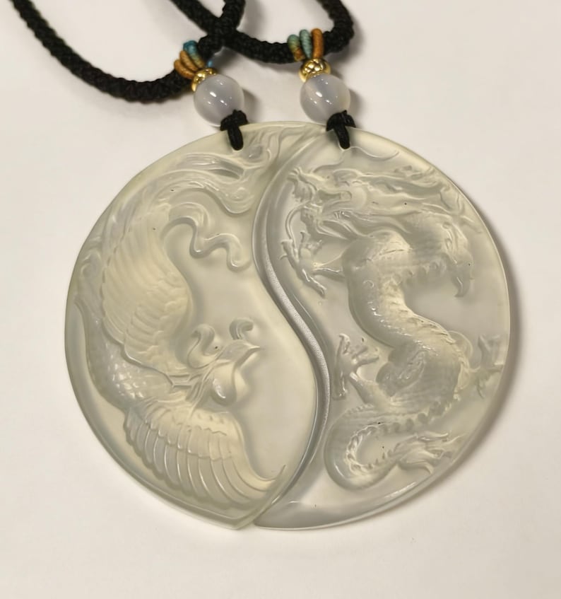 May include: A pair of light-colored pendants, each in a half-circle shape, forming a Yin and Yang symbol. One pendant features a carved dragon, the other a phoenix. The pendants are connected to black cords with decorative beads.