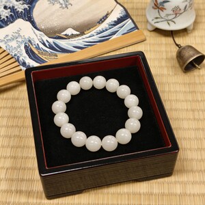 May include: A white beaded bracelet displayed in a black and red lacquered box. The bracelet is composed of round, opaque beads. A Japanese fan with a wave design and a small bell are in the background.