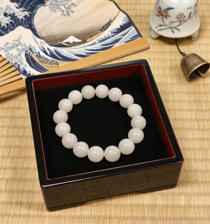 May include: A white beaded bracelet displayed in a black and red lacquered box. The bracelet is composed of round, opaque beads. A Japanese fan with a wave design and a small bell are in the background.