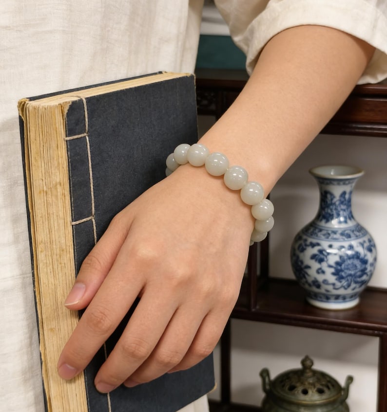 May include: A light green beaded bracelet with large, round beads is worn on a wrist. The person is holding a dark blue book with aged, yellowed pages. A blue and white vase is in the background.