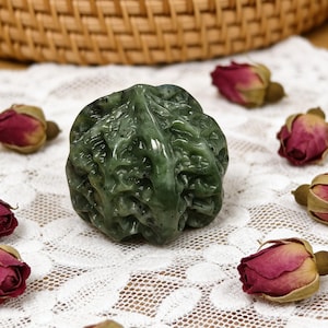 Pode incluir: Uma escultura de jade verde com uma superfície texturizada, assemelhando-se a uma formação rochosa natural. A escultura é cercada por rosas vermelhas secas em um tecido de renda branco. Uma cesta tecida está ao fundo.