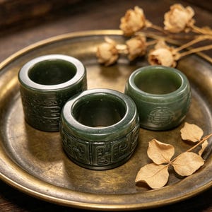 May include: Three dark green jade rings with carved patterns sit on a gold-colored metal tray. The rings have a cylindrical shape with detailed carvings. Dried flowers and leaves are arranged around the rings, creating a natural aesthetic.