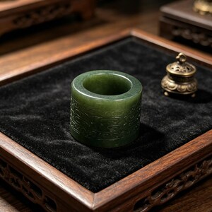 May include: A dark green jade ring with carved decorative patterns. The ring is cylindrical with a smooth, polished surface. It sits on a black velvet surface within a wooden tray. A small, ornate metal incense burner is in the background, suggesting a display setting.