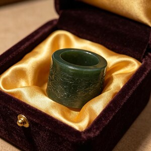 May include: A dark green jade ring with detailed carvings, displayed in a burgundy velvet box with a gold satin lining. The ring features intricate patterns, demonstrating artistry and craftsmanship. The box is secured with a small gold clasp.